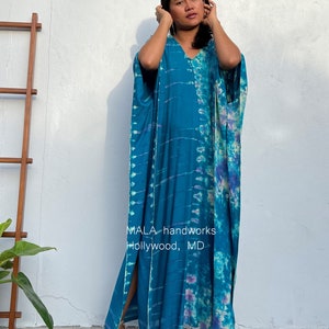May include: A long, teal blue kaftan with a tie-dye pattern in shades of blue, purple, and white. The kaftan has a V-neck and side slits. The text "MALA handworks Hollywood, MD" is printed on the front of the kaftan.