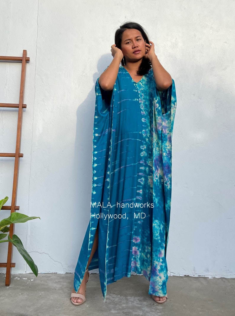May include: A long, teal blue kaftan with a tie-dye pattern in shades of blue, purple, and white. The kaftan has a V-neck and side slits. The text "MALA handworks Hollywood, MD" is printed on the front of the kaftan.