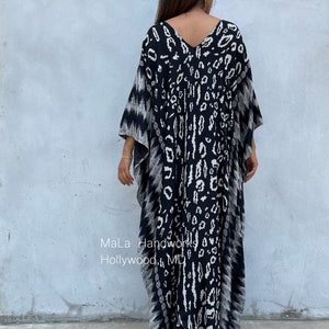 May include: Black and white kaftan with a leopard print design. The kaftan has a V-neck and long sleeves. The model is wearing the kaftan with a pair of nude heels. The text "MaLa Handworks Hollywood, MD" is visible on the bottom of the kaftan.