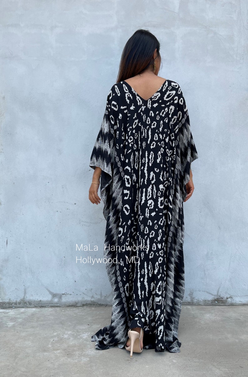 May include: Black and white kaftan with a leopard print design. The kaftan has a V-neck and long sleeves. The model is wearing the kaftan with a pair of nude heels. The text "MaLa Handworks Hollywood, MD" is visible on the bottom of the kaftan.