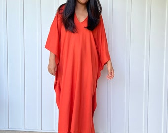 Puede incluir: Un vestido caftán de color naranja-rojo con cuello en V. El vestido tiene mangas anchas y llega hasta los tobillos. La persona lleva sandalias marrones con correas. El fondo es una pared blanca con paneles verticales.