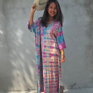Op de afbeelding: Een vrouw draagt een lange, roze, blauwe en witte tie-dye jurk met een witte strohoed. Ze staat op een betonnen oppervlak.