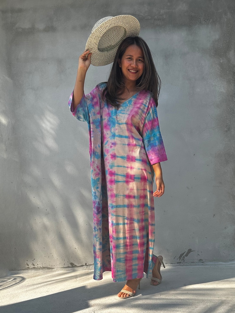 Op de afbeelding: Een vrouw draagt een lange, roze, blauwe en witte tie-dye jurk met een witte strohoed. Ze staat op een betonnen oppervlak.