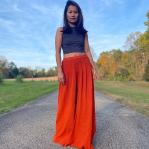 May include: A woman wearing a grey sleeveless top and wide-legged orange trousers. She is standing on a paved road with green grass on either side.