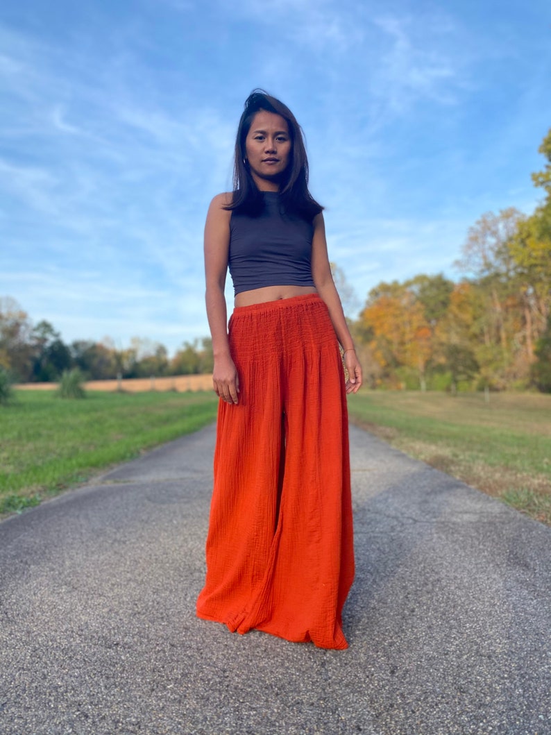 May include: A woman wearing a grey sleeveless top and wide-legged orange trousers. She is standing on a paved road with green grass on either side.