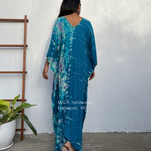 May include: A long, flowing, teal blue and purple tie-dye kaftan with a V-neckline. The kaftan is made of a soft, lightweight fabric and has a relaxed fit. The model is wearing the kaftan with a pair of nude heels. The text "MALA handworks Hollywood, MD" is visible on the bottom of the kaftan.