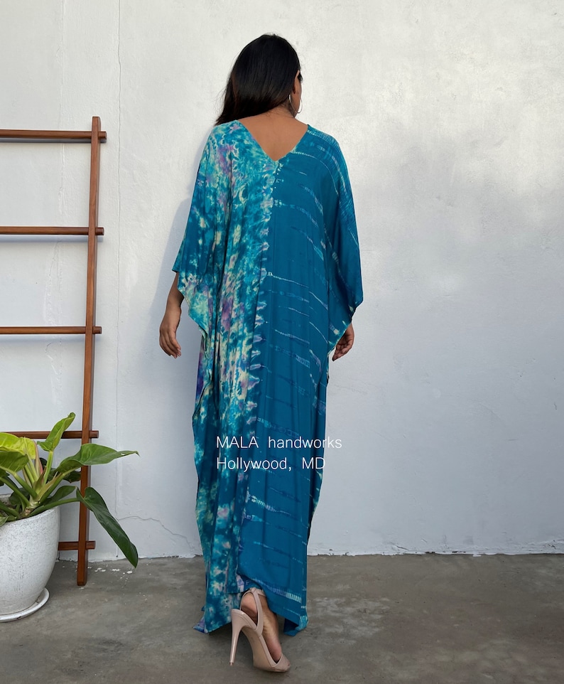 May include: A long, flowing, teal blue and purple tie-dye kaftan with a V-neckline. The kaftan is made of a soft, lightweight fabric and has a relaxed fit. The model is wearing the kaftan with a pair of nude heels. The text "MALA handworks Hollywood, MD" is visible on the bottom of the kaftan.
