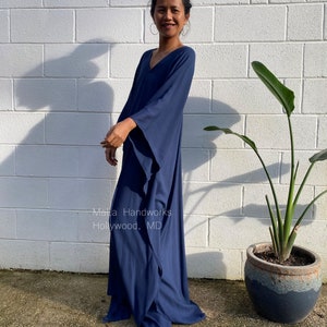 May include: A woman wearing a long, navy blue, flowing kaftan dress with a V-neck and long sleeves. The dress has a relaxed fit and is made of a soft, comfortable fabric. The woman is standing in front of a white brick wall with a green plant in a pot to the right. The text "Malta Handworks Hollywood, MD" is visible on the dress.