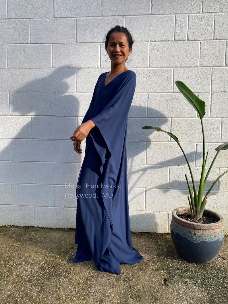 May include: A woman wearing a long, navy blue, flowing kaftan dress with a V-neck and long sleeves. The dress has a relaxed fit and is made of a soft, comfortable fabric. The woman is standing in front of a white brick wall with a green plant in a pot to the right. The text "Malta Handworks Hollywood, MD" is visible on the dress.
