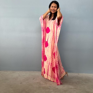 May include: A long, flowing, tie-dye kaftan in shades of pink and cream. The dress features large, circular pink accents and vertical pink stripes. The garment has wide sleeves and a V-neckline. The model is wearing high heels.