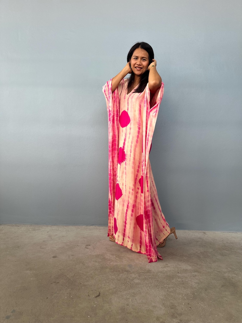 May include: A long, flowing, tie-dye kaftan in shades of pink and cream. The dress features large, circular pink accents and vertical pink stripes. The garment has wide sleeves and a V-neckline. The model is wearing high heels.