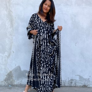 May include: A black and white patterned maxi dress with a V-neckline and long sleeves. The dress has a tie at the waist and is made of a soft, flowing fabric. The dress is being worn by a person standing in front of a gray wall. The text "Mala Handworks Hollywood, MD" is visible on the dress.