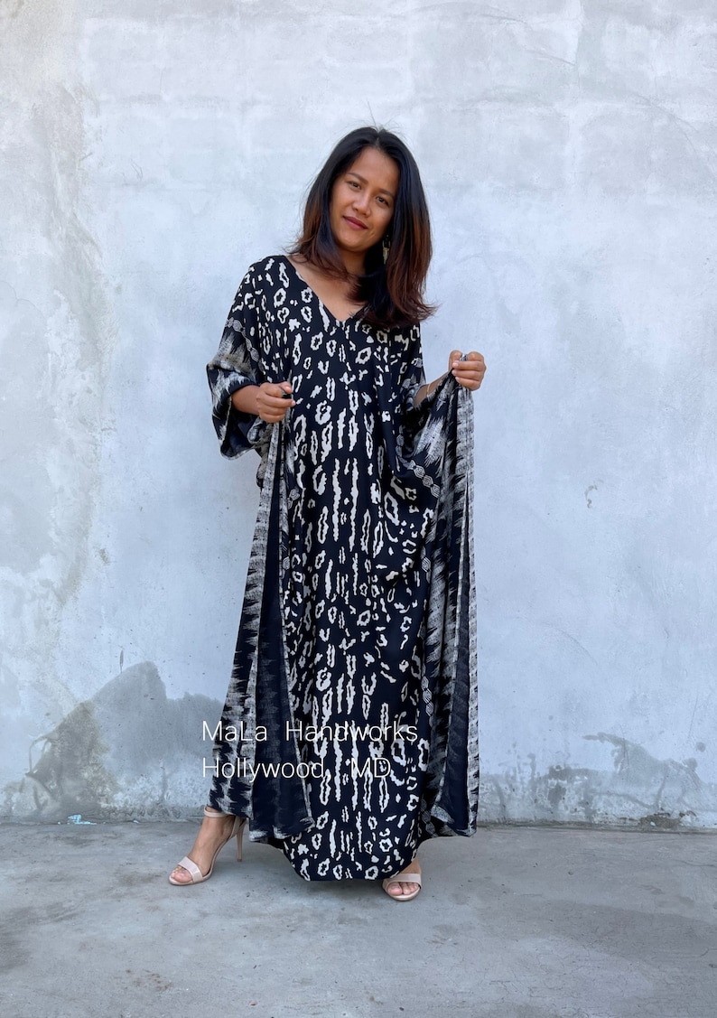 May include: A black and white patterned maxi dress with a V-neckline and long sleeves. The dress has a tie at the waist and is made of a soft, flowing fabric. The dress is being worn by a person standing in front of a gray wall. The text "Mala Handworks Hollywood, MD" is visible on the dress.