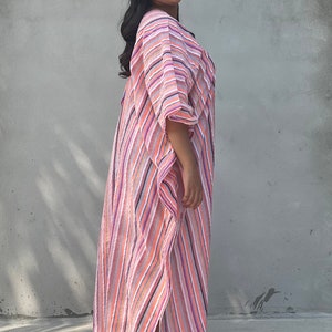 May include: A woman wearing a long, pink and white striped dress with a flowing silhouette. The dress has a relaxed fit and a V-neckline. She is standing in front of a gray wall.