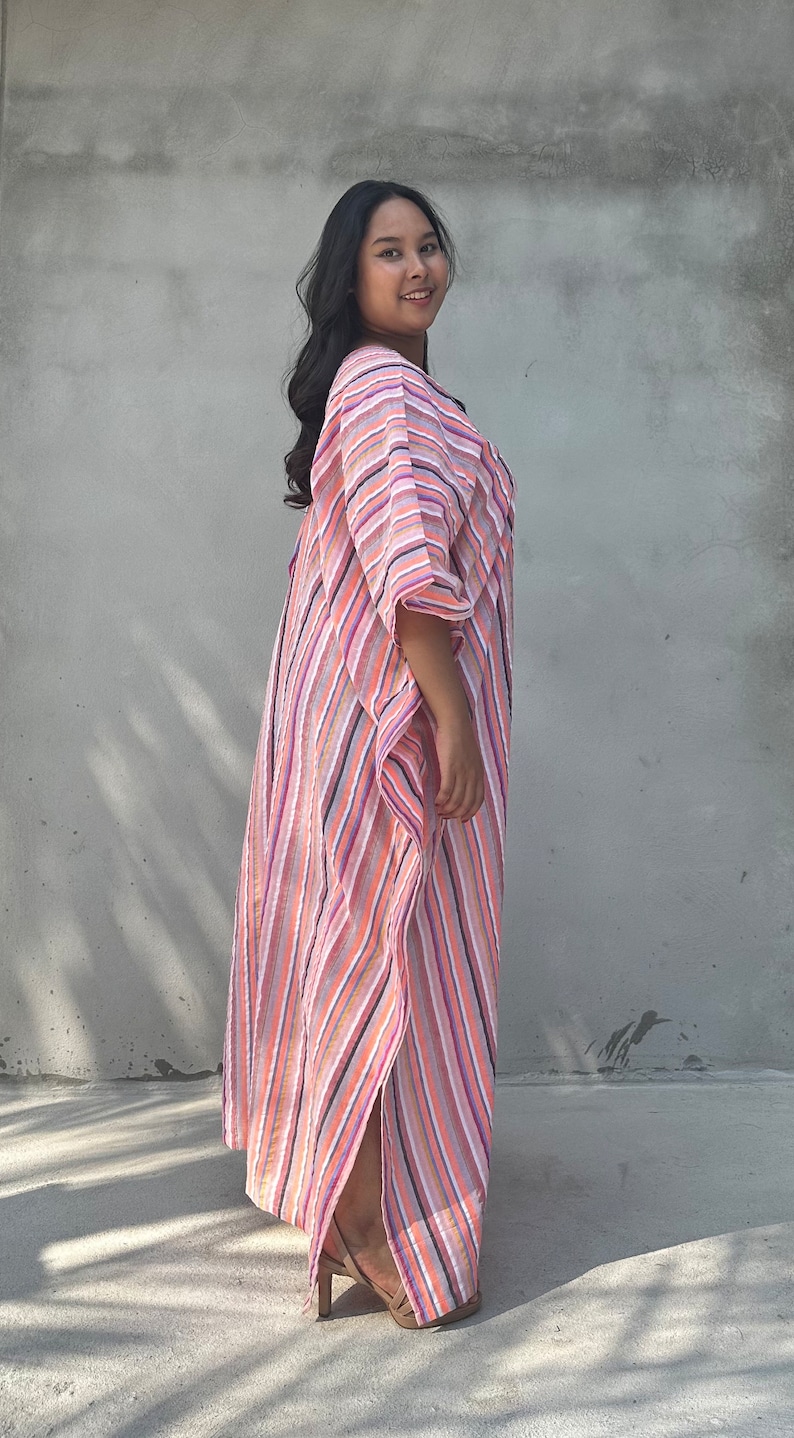 May include: A woman wearing a long, pink and white striped dress with a flowing silhouette. The dress has a relaxed fit and a V-neckline. She is standing in front of a gray wall.