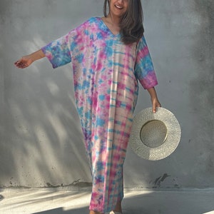Op de afbeelding: Een vrouw draagt een lange, losse tie-dye jurk in tinten roze, blauw en wit. Ze glimlacht en houdt een grote, witte strohoed vast.