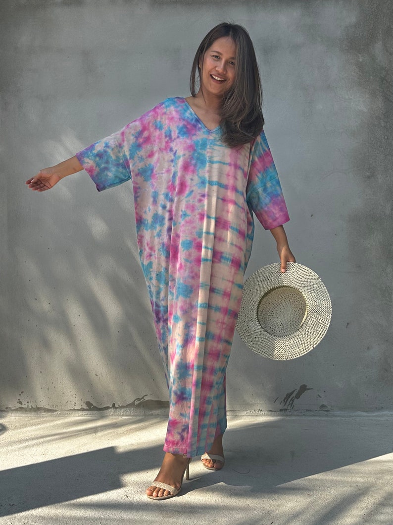 Op de afbeelding: Een vrouw draagt een lange, losse tie-dye jurk in tinten roze, blauw en wit. Ze glimlacht en houdt een grote, witte strohoed vast.