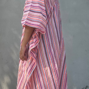 May include: A woman wearing a long, pink and white striped kaftan with a diagonal pattern. The kaftan has a loose fit and is made of a lightweight fabric.