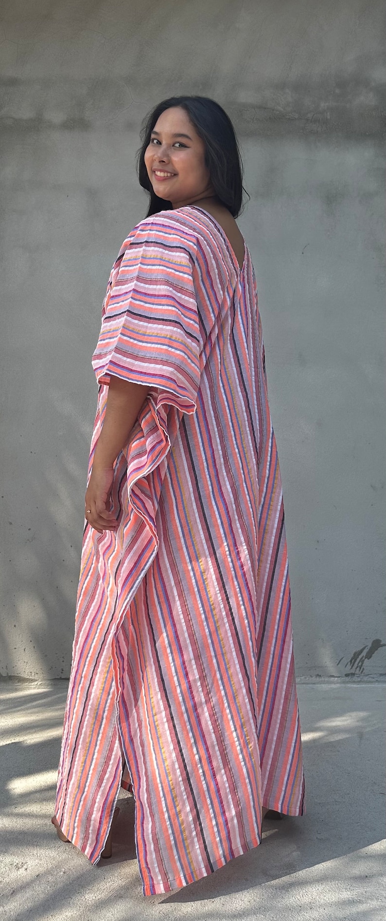 May include: A woman wearing a long, pink and white striped kaftan with a diagonal pattern. The kaftan has a loose fit and is made of a lightweight fabric.