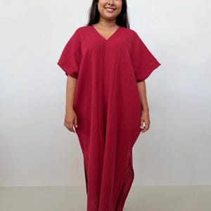 Può includere: Un abito caftano rosso con scollo a V e maniche corte. L'abito è realizzato in un tessuto strutturato e presenta spacchi laterali. La modella sorride e indossa l'abito davanti a uno sfondo bianco.