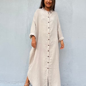 May include: A long, beige linen shirt dress with a button-up front and long sleeves. The dress has a relaxed fit and a slightly curved hemline.