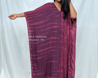 Puede incluir: Un vestido largo y fluido de caftán burdeos con cuello en V. El vestido presenta un estampado tie-dye con rayas horizontales. El texto "MALA handworks Hollywood, MD" es visible. El vestido está hecho de una tela suave y fluida.