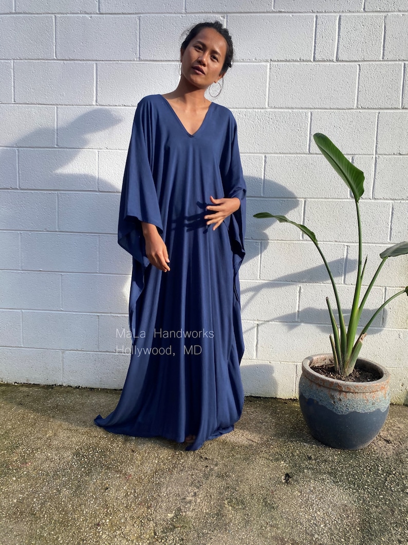 May include: A navy blue maxi dress with a V-neck and long sleeves. The dress is made of a soft, flowing fabric and has a relaxed fit. The dress is shown being worn by a person standing in front of a white brick wall. The text "MaLa Handworks Hollywood, MD" is visible on the dress.