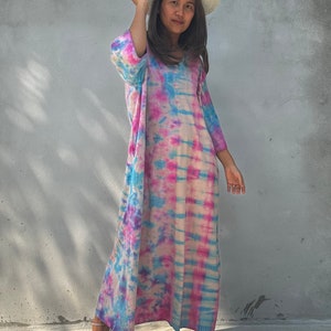 Op de afbeelding: Een vrouw draagt een lange, roze, blauwe en witte tie-dye jurk met een strohoed. Ze staat voor een grijze muur.
