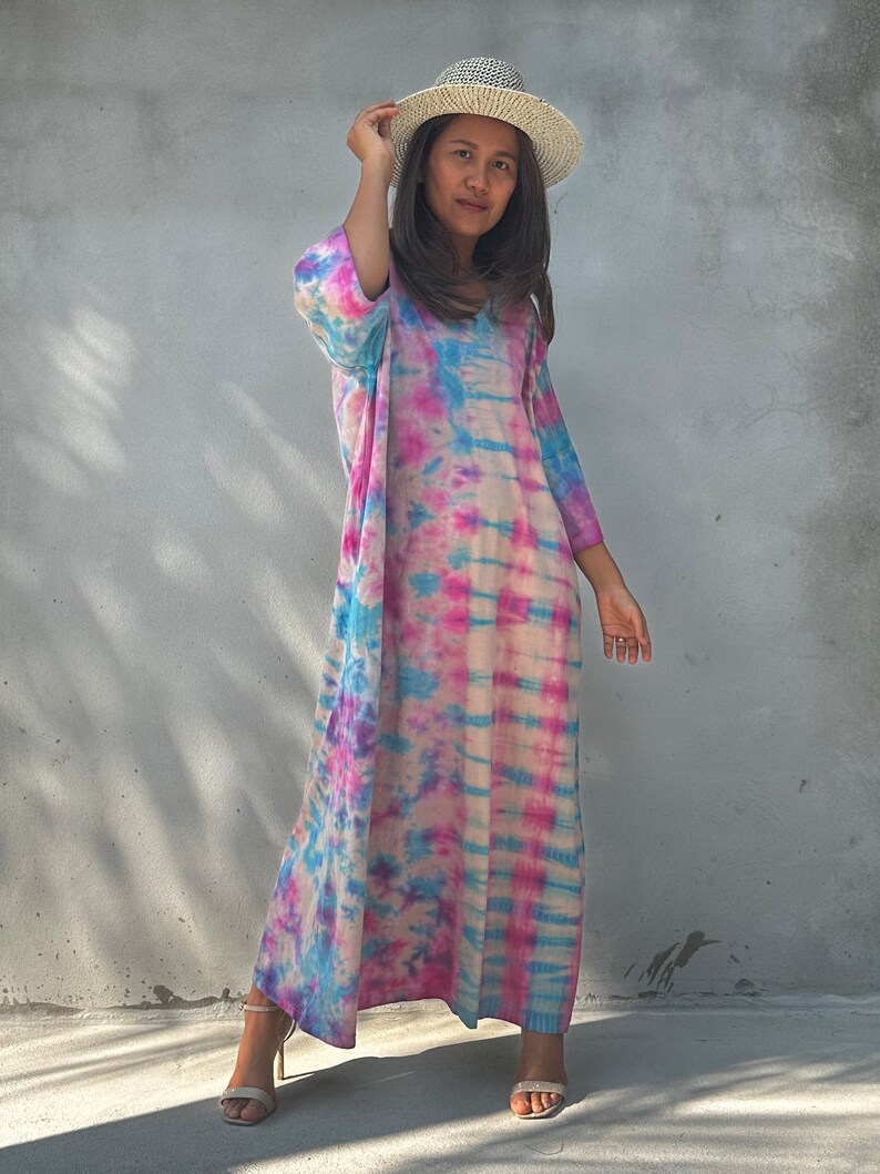 Op de afbeelding: Een vrouw draagt een lange, roze, blauwe en witte tie-dye jurk met een strohoed. Ze staat voor een grijze muur.