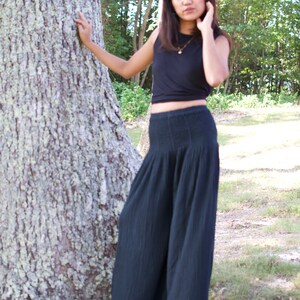 May include: A woman wearing a black crop top and wide-legged black pants stands in a forest setting. She is leaning against a large tree trunk.