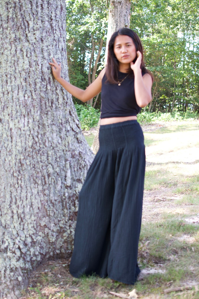 May include: A woman wearing a black crop top and wide-legged black pants stands in a forest setting. She is leaning against a large tree trunk.