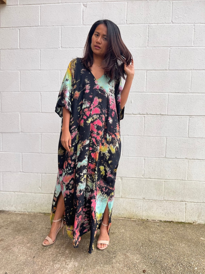May include: A long, black maxi dress with a tie-dye pattern in shades of pink, blue, and yellow. The dress has a V-neck and short sleeves. It features side slits and is made of a soft, flowing fabric.