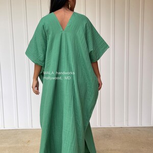 May include: A green caftan dress with a V-neck and wide sleeves. The dress is made of a lightweight fabric and has a loose, flowing fit. The text "MALA handworks Hollywood, MD" is visible on the dress.