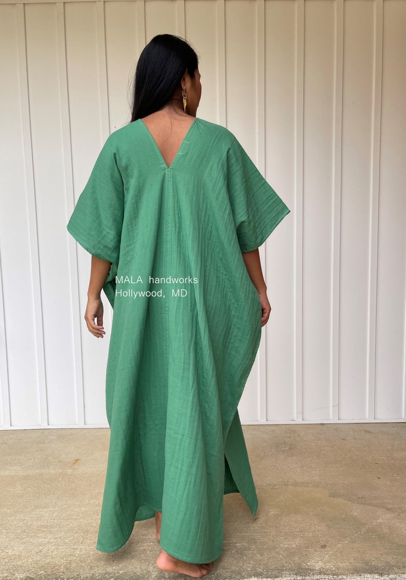 May include: A green caftan dress with a V-neck and wide sleeves. The dress is made of a lightweight fabric and has a loose, flowing fit. The text "MALA handworks Hollywood, MD" is visible on the dress.