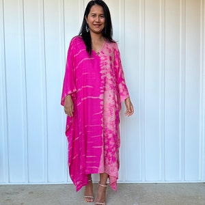 May include: A vibrant pink tie-dye caftan dress with a V-neck and wide sleeves. The dress is split vertically, with one side a solid bright pink and the other a lighter pink tie-dye pattern. The model is wearing silver earrings and strappy sandals.