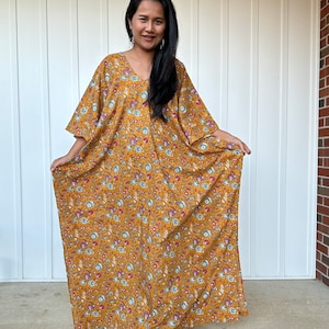 May include: A long, flowing mustard yellow kaftan with a floral print. The kaftan has a V-neck and wide sleeves. The model is wearing the kaftan with bare feet.