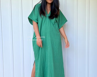 May include: A woman wearing a long, green, loose-fitting dress with a V-neck and short sleeves. The dress has a side slit and is made of a textured fabric.