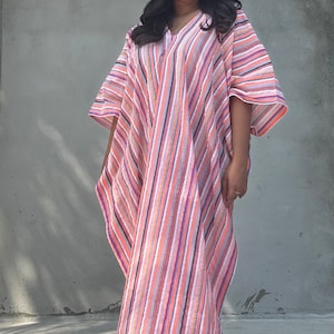 May include: A woman wearing a long, pink and white striped kaftan with a V-neckline. The kaftan has a loose, flowing fit and is made of a lightweight fabric.