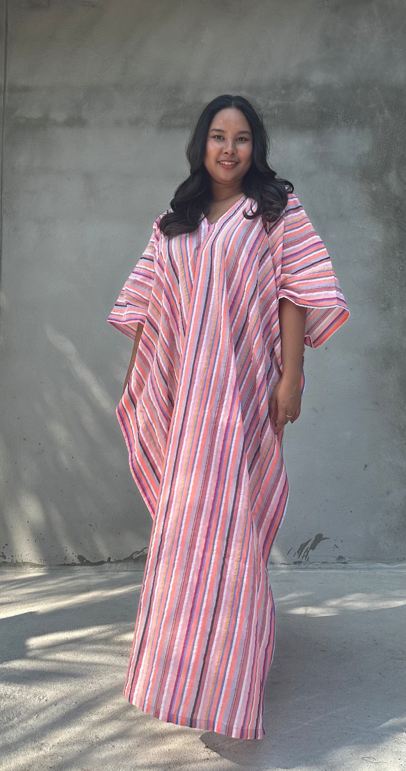 May include: A woman wearing a long, pink and white striped kaftan with a V-neckline. The kaftan has a loose, flowing fit and is made of a lightweight fabric.