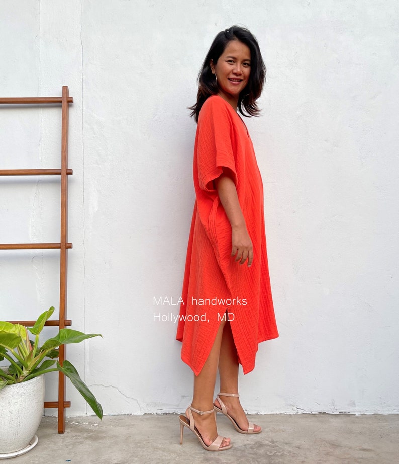 May include: A woman wearing a bright orange, loose-fitting, knee-length dress with a V-neck and short sleeves. The dress has a slightly asymmetrical hemline. The woman is standing on a tiled surface with a white wall and a wooden ladder in the background. The text "MALA handworks Hollywood, MD" is visible on the dress.