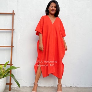 May include: A woman wearing a bright orange, loose-fitting, knee-length dress with a V-neck and short sleeves. The dress has a side slit and is made of a textured fabric. The text "MALA handworks Hollywood, MD" is visible on the dress.