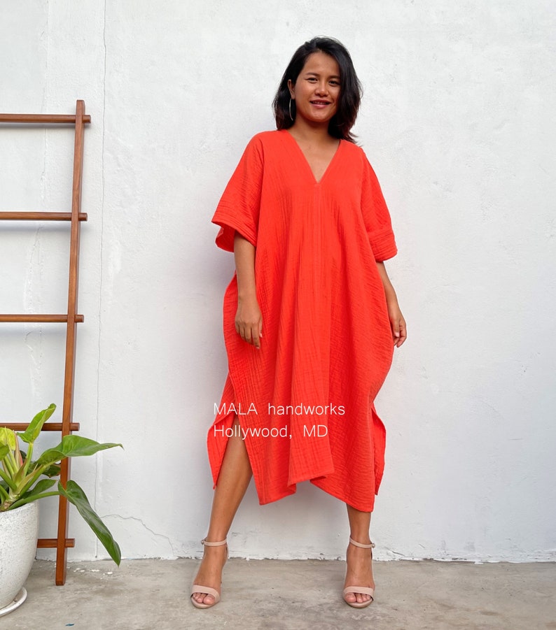May include: A woman wearing a bright orange, loose-fitting, knee-length dress with a V-neck and short sleeves. The dress has a side slit and is made of a textured fabric. The text "MALA handworks Hollywood, MD" is visible on the dress.