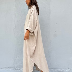 May include: A long, loose-fitting, beige linen dress with a button-up front and long sleeves. The dress has a relaxed, casual fit and is perfect for warm weather.