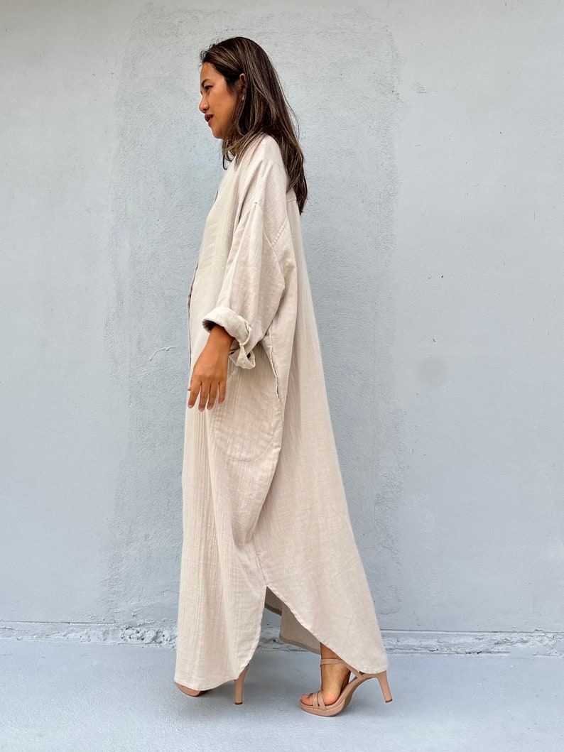 May include: A long, loose-fitting, beige linen dress with a button-up front and long sleeves. The dress has a relaxed, casual fit and is perfect for warm weather.