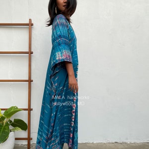 May include: A woman wearing a long, flowing teal blue and pink tie-dye maxi dress with a V-neck and 3/4 sleeves. The dress has a flowing, asymmetrical hemline. The dress is made by MALA handworks, Hollywood, MD.