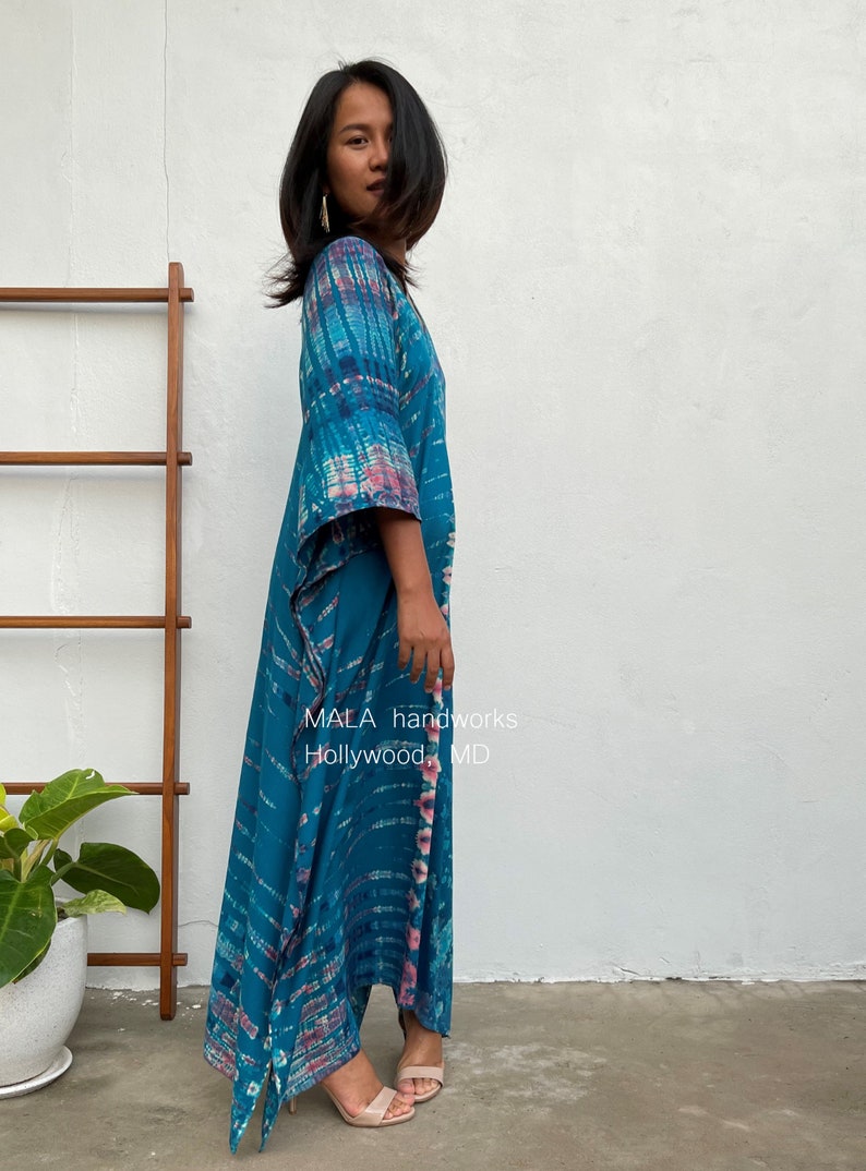 May include: A woman wearing a long, flowing teal blue and pink tie-dye maxi dress with a V-neck and 3/4 sleeves. The dress has a flowing, asymmetrical hemline. The dress is made by MALA handworks, Hollywood, MD.