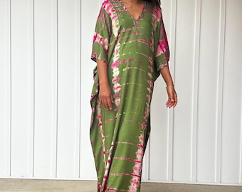 Op de afbeelding: Een lange, vloeiende groene jurk met een V-hals en korte mouwen. De jurk heeft een tie-dye patroon in roze en witte strepen. De vrouw draagt beige sandalen.