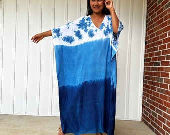 May include: A woman wearing a long, blue and white tie-dye kaftan dress with a V-neck. She is standing on a concrete porch with a brick wall in the background.