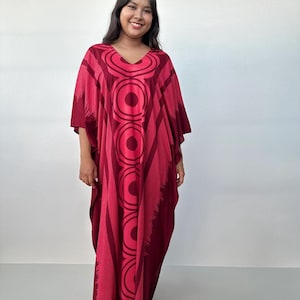 May include: A floor-length kaftan dress in shades of deep burgundy and bright magenta. The garment features a V-neckline and a prominent vertical pattern of concentric circles running down the center, complemented by soft, brush-like side accents.