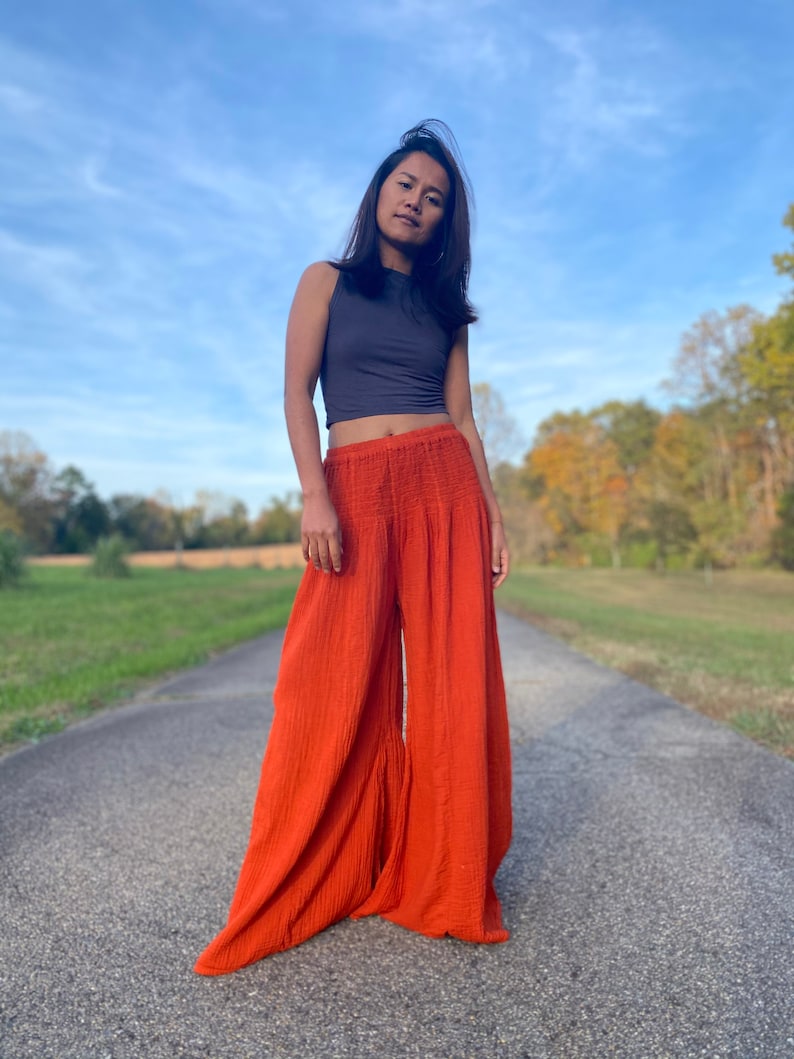 May include: A woman wearing a grey sleeveless top and wide-legged, burnt orange trousers. She is standing on a paved road with a grassy field and trees in the background.